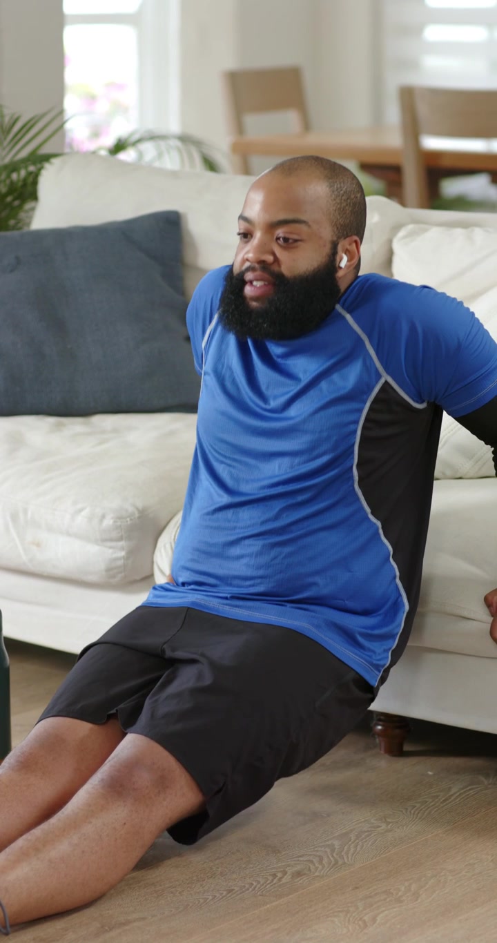 Vertical video: Adult African man bracing on sofa edge, doing triceps dips at home for strength