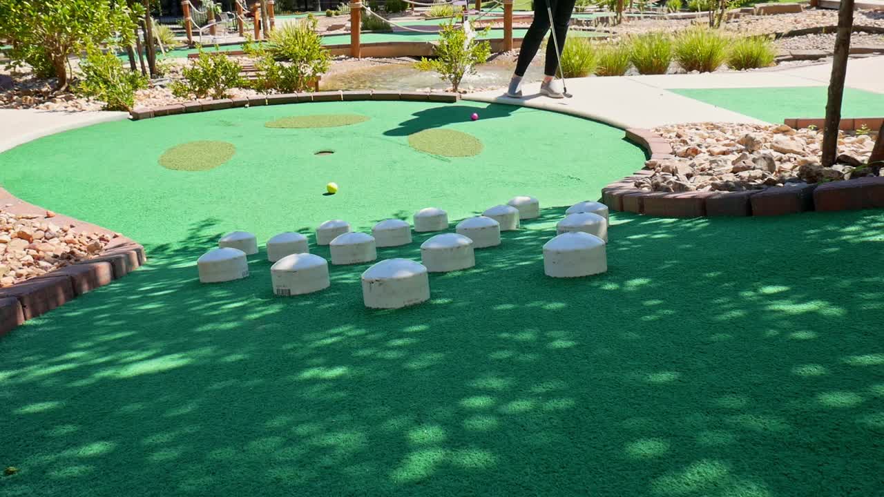 Couple playing mini golf and hitting a yellow golf ball between white post obstacles on green astroturf
