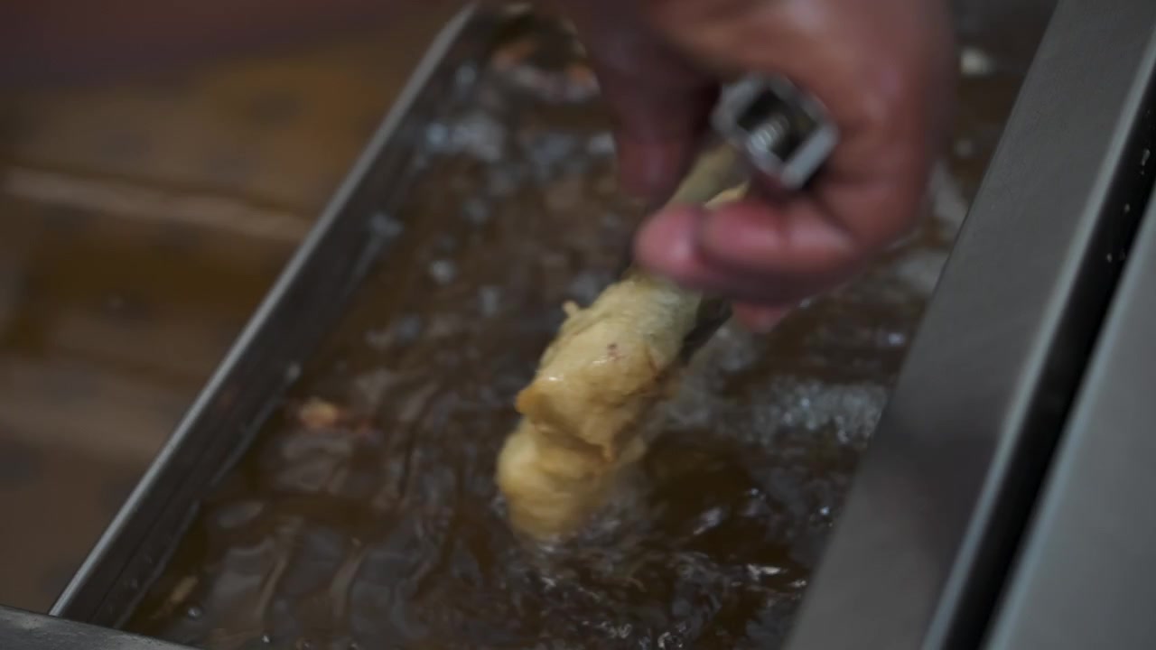 Coated fish fillet is lowered into bubbling hot oil with a utensil as it begins to fry in a store fried foods setup.