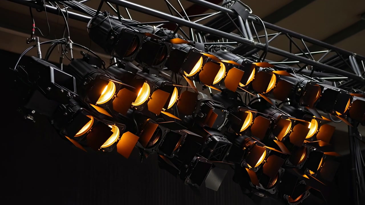 Dense array of black spotlights mounted to steel truss inside performance venue, with more lamps glowing warm amber and creating strong contrast, metallic texture, and technical atmosphere