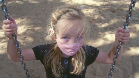 Cute-Young-Girl-With-Face-Mask-Looking-At-The-Camera-While-Sitting-On-The-Swing-At-The-Playground-On-A-Sunny-Morning---high-angle,-close-up