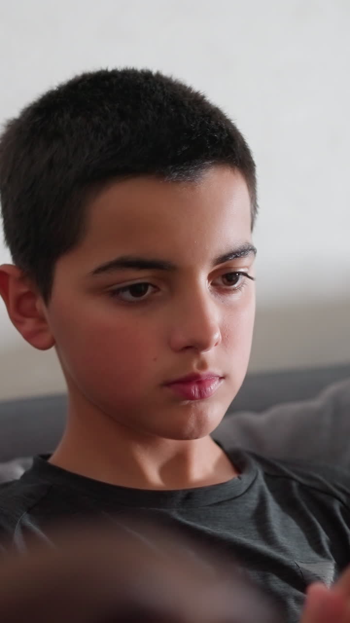 Niño reflexivo leyendo con gran concentración en el sofá y hojeando las páginas de un libro en un entorno interior tranquilo mientras una mujer parcialmente visible se sienta cerca