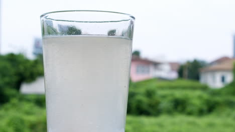agua burbujeante en un vaso causada por la dilución de un antiácido o bicarbonato de sodio en un telón de fondo de casas y vegetación verde fuera de foco