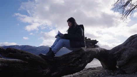young woman sits on a large piece of driftwood reading a book, framed by cloudy skies, mountain silhouettes, and the peaceful patagonian lakeshore, real time, static camera