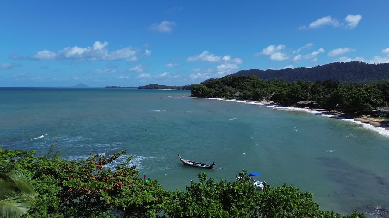 Aerial drone flight over green palm trees and tropical jungle on Koh Lanta, Thailand, opening to a secluded white sandy beach and turquoise ocean with boats, mountains, and peaceful island scenery