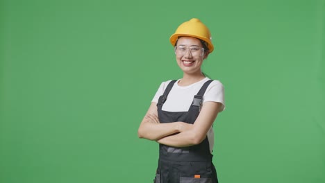 side view of asian woman worker wearing goggles and safety helmet smiling and crossing her arms while standing in the green screen background studio