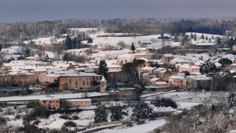 Pequeño-Pueblo-Bajo-La-Nieve-En-Francia,-Monpazier-En-Dordoña,-La-Mañana-Se-Levanta-Sobre-El-Campo,-Un-Drone-Circular-Rodó-Alrededor-De-La-Iglesia-Iluminada-Por-El-Sol-De-La-Mañana