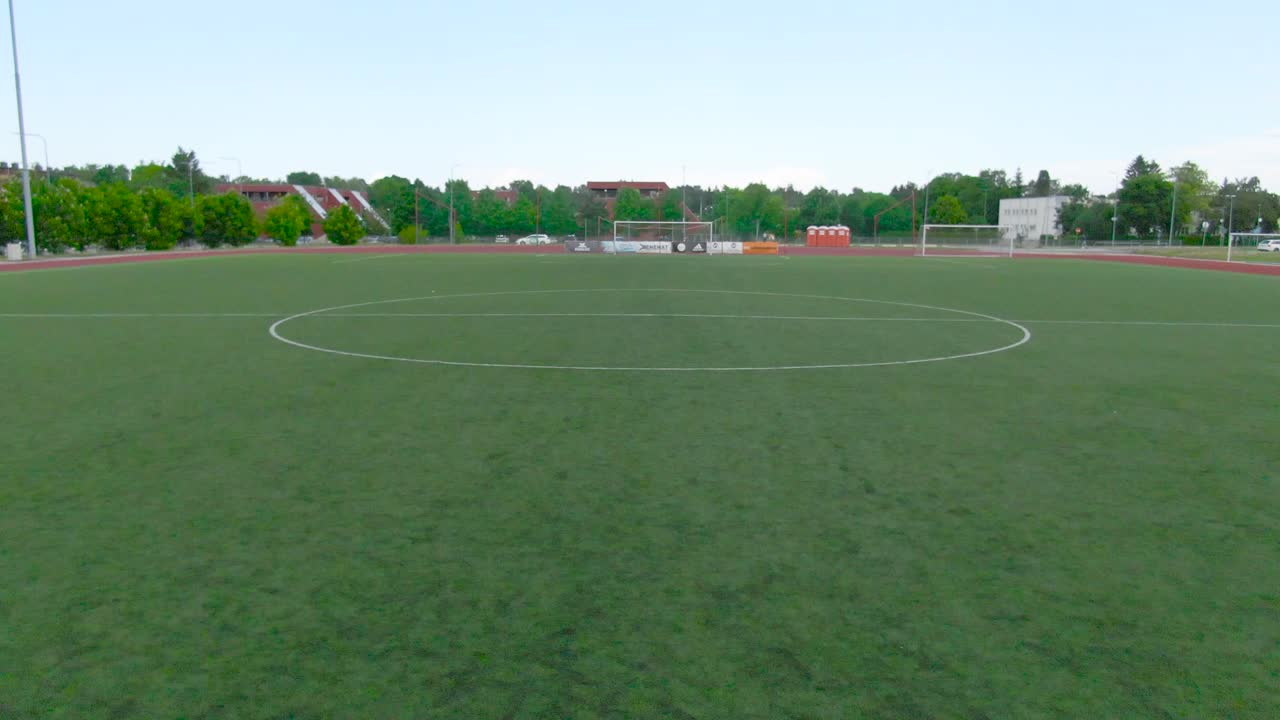 Aerial drone footage fly over from Saue Laagri soccer or football stadium during cloudy weather summer or autumn time. Green trees, asphalt roads, cars and houses in the background. Cars driving too.