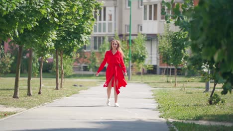 backside view of long-haired woman outdoors. pretty girl in red dress walking along the street and turns to camera with a smile. slow motion.