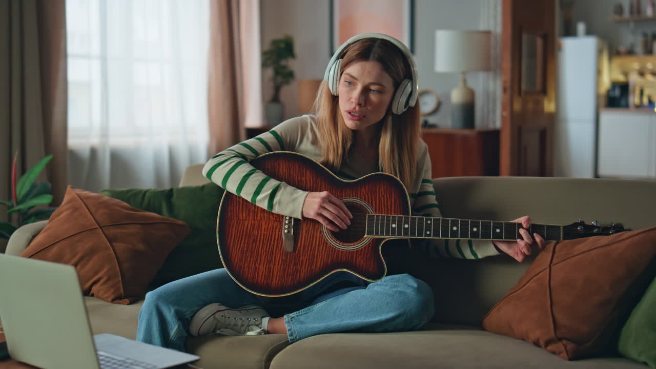 Gitarristkvinna med hörlurar komponerar musik på akustisk gitarr i soffan hemma.
