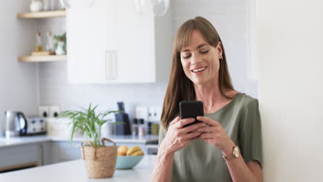 Una-Mujer-Caucásica-De-Mediana-Edad-Envía-Mensajes-De-Texto-A-Su-Teléfono-En-Una-Cocina-Moderna