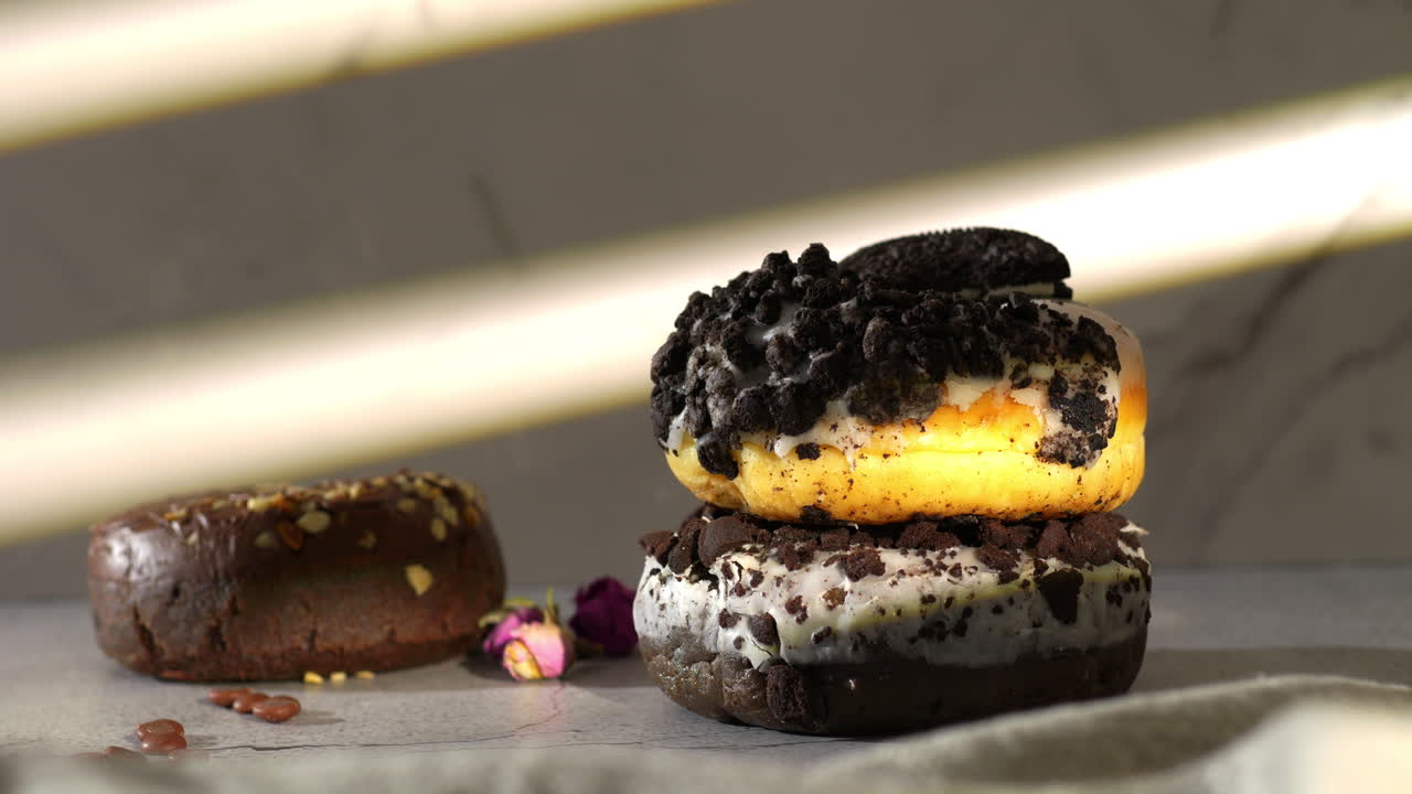 A hand with pink nails reaches to take a white-glazed donut topped with cookie crumbles from a stack. warmly lit by natural sunlight with another chocolate donut and rosebuds nearby on a moody surface