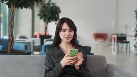 young-and-beautiful-woman-in-a-suit-sits-on-the-couch-in-the-business-center,-enthusiastically-playing-a-mobile-game-on-her-smartphone-with-a-smile,-then-angrily-throws-the-phone