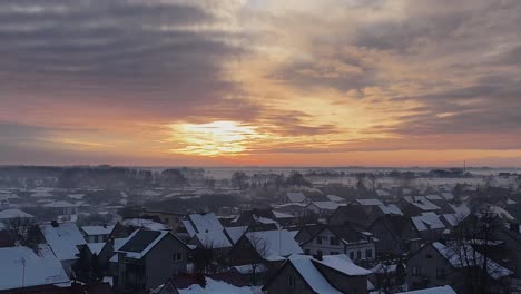 aerial pullback above snow covered suburban roofs with fire yellow sunset streak in sky above misty fog