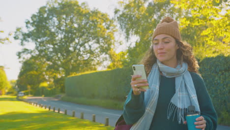 Mujer-Joven-Sonriente-Con-Sombrero-Y-Bufanda-Caminando-En-El-Campo-De-Otoño-Mirando-El-Teléfono-Móvil