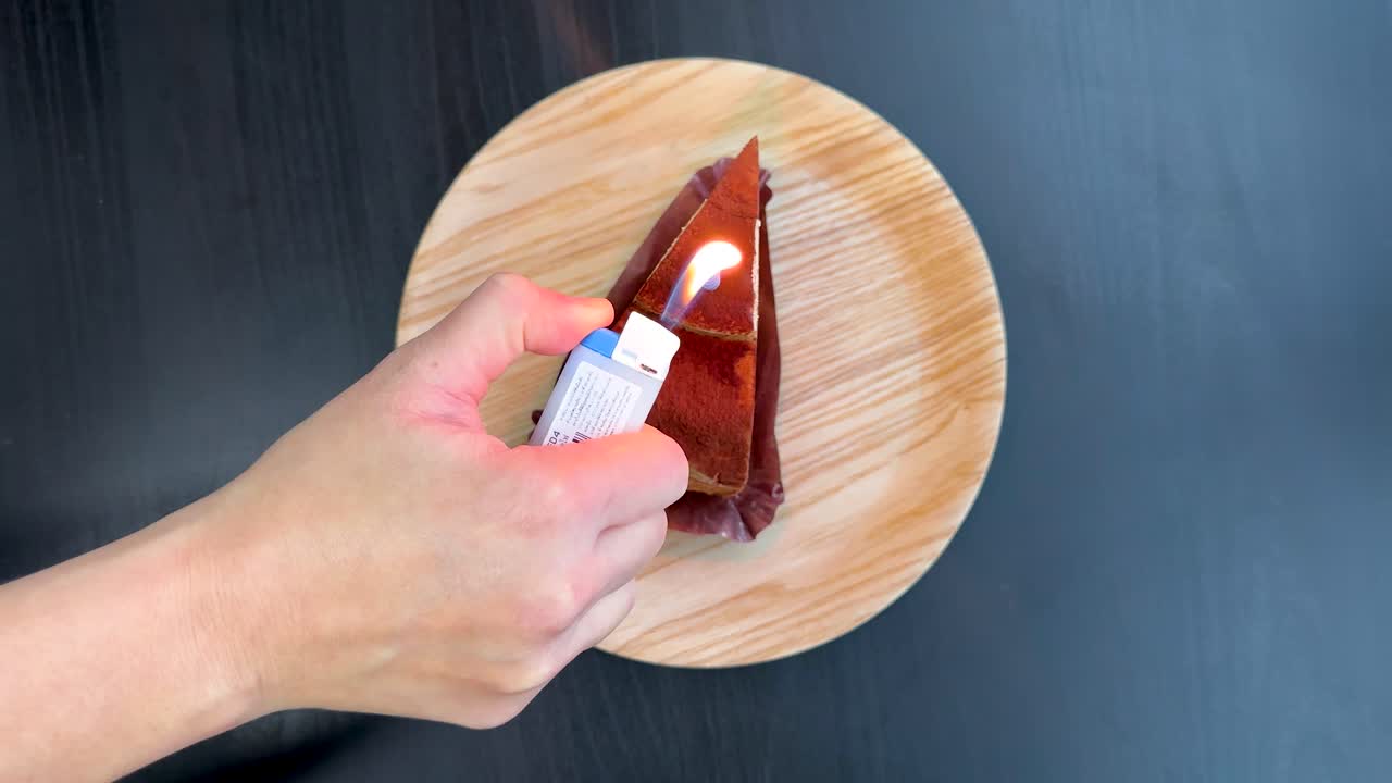 Hand lights single candle on tiramisu cake slice, overhead view, bright even lighting, steady shot