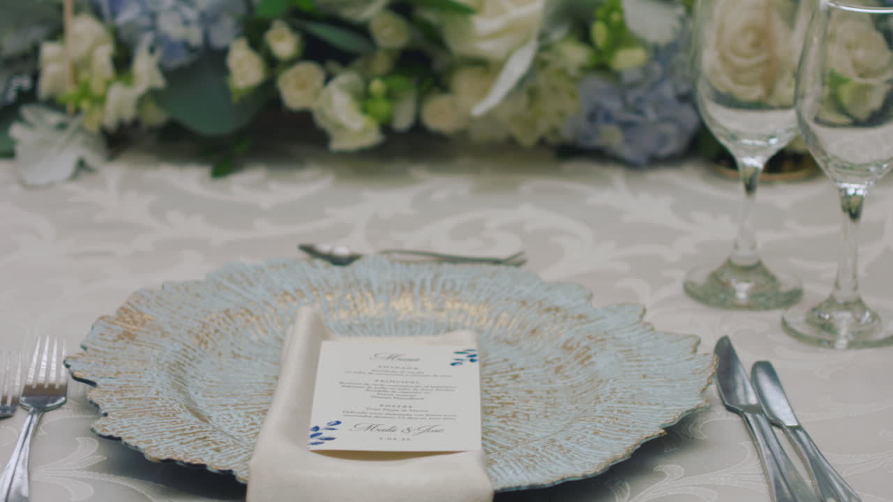 A detailed clip of an elegantly decorated wedding table. It features stemware, a white tablecloth, plates, cutlery, cloth napkins, a delicate floral arrangement, and a personalized invitation.