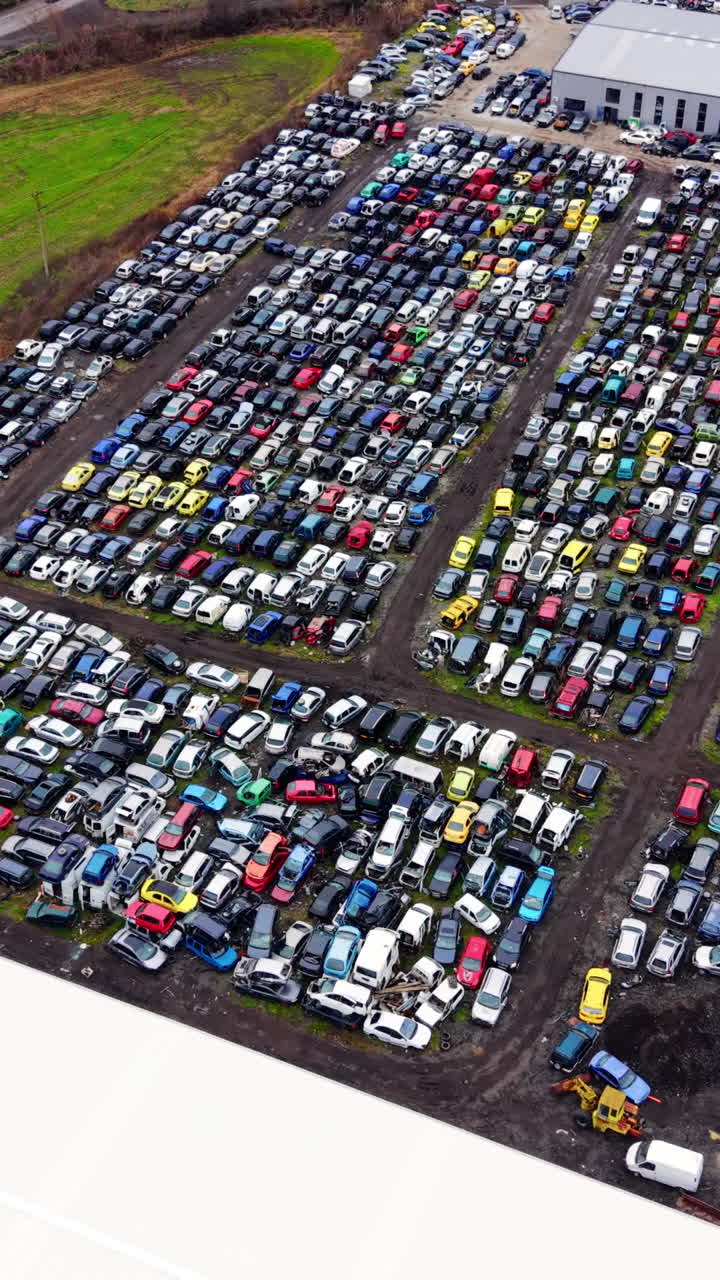 Filas organizadas de coches desguazados y usados en una planta de reciclaje junto a un edificio industrial. Vista aérea de un gran desguace de coches al aire libre con filas de vehículos. Vídeo vertical