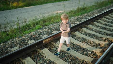 aerial view of young boy walking along rail track in casual clothes, dragging foliage in hand while surrounded by gravel, wild grass, and unpaved rural path under soft daylight