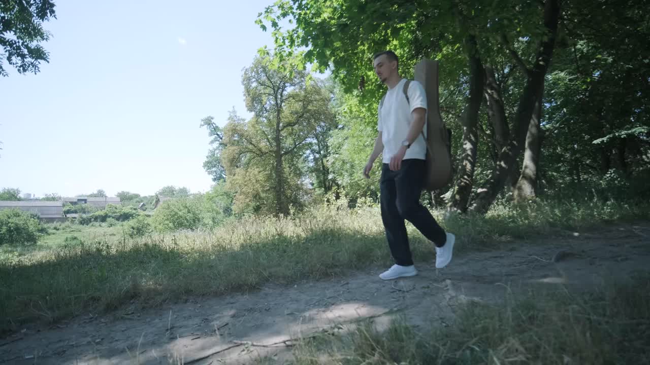 Mladý muž jde s kytarou po ulici u lesa