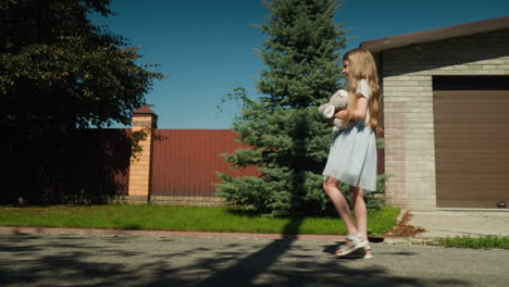 Young girl in blue dress walking with stuffed bunny under arm along paved suburban street as shadow from tree and pole falls across ground near house with brown garage and brick wall