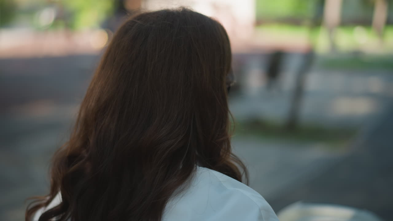 Wanita menikmati kopi. Gadis di taman menyeruput kopi dengan tenang. Tampilan belakang wanita yang tenggelam dalam momen kopi yang hening. Wanita Kaukasia dengan rambut tergerai merenung sendirian di jalan setapak taman sambil minum kopi