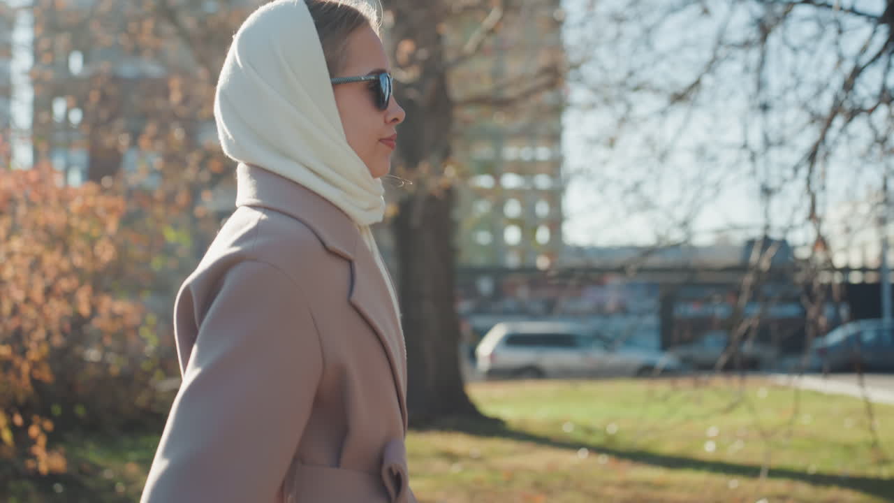 Elegante vrouw wandelt in het park met ivoorkleurige hoofddoek en beige jas, zonnebril, zacht tegenlicht, gouden bladertooi, stedelijke skyline, contemplatieve stemming, slow motion close-ups, fashion blogger styling.