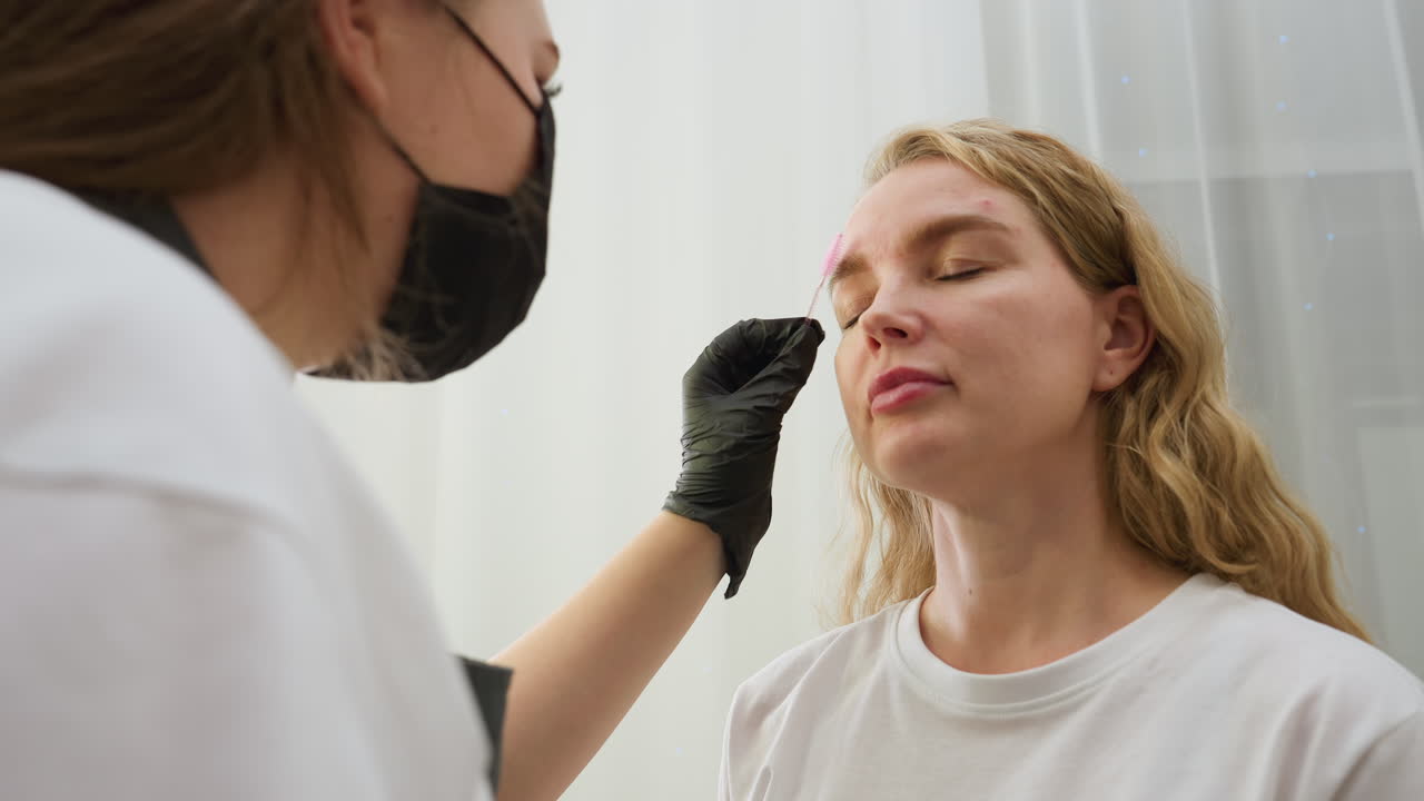 Beautician wearing gloves carefully applies substance onto client eyebrow using small brush while client looks upward in bright indoor salon with white curtains and relaxed atmosphere