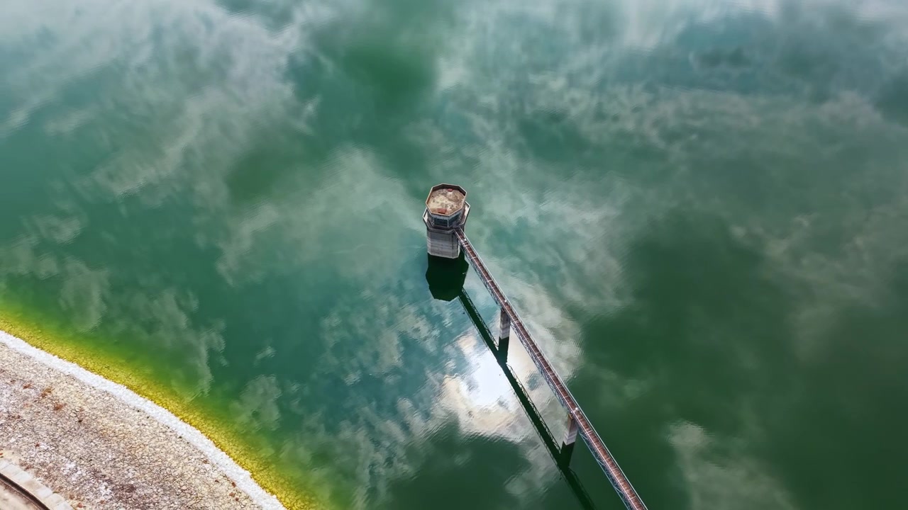 Water tower with a clear reflection in a calm lake during the day near a shore with stones and green algae