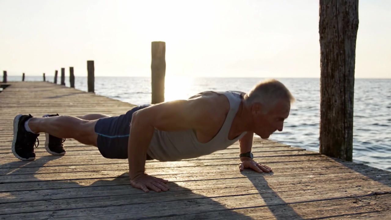 Un Uomo Anziano Determinato Esegue Flessioni su un Molo di Legno Mentre il Sole Tramonta su Acque Calme, Dimostrando Forza e Perseveranza a Qualsiasi Età
