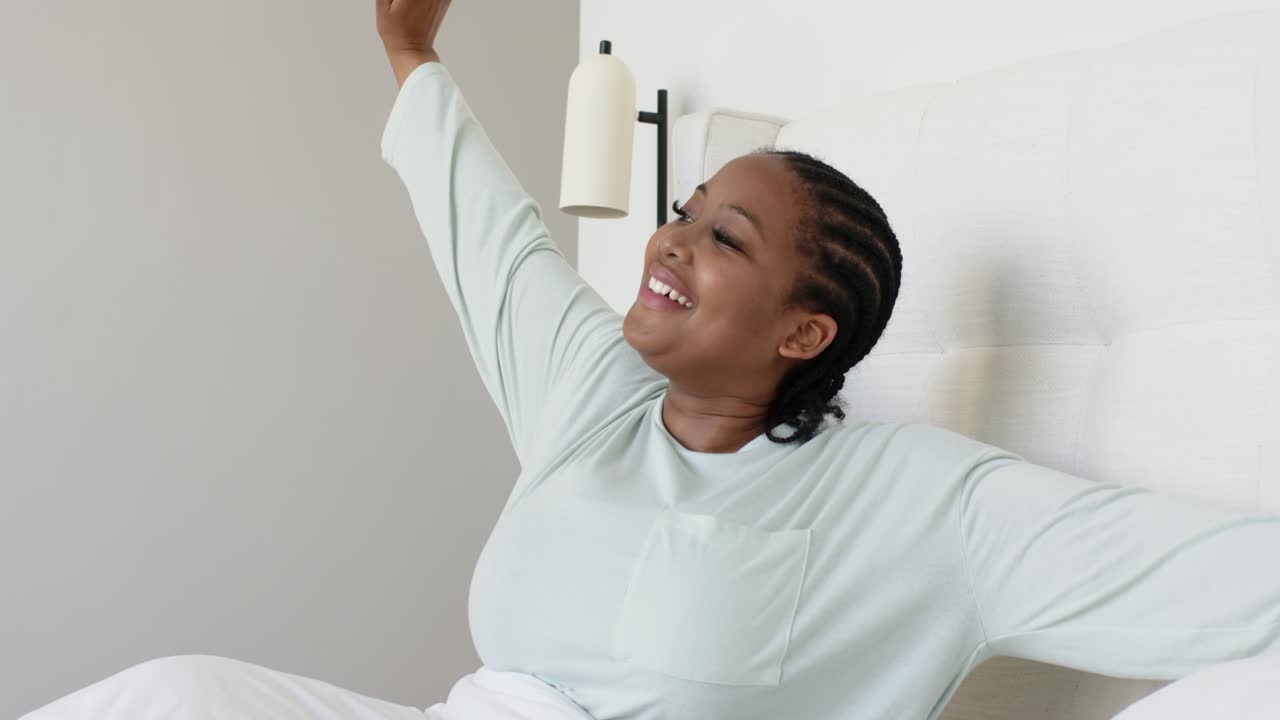 Donna afroamericana di mezza età che si sveglia nel letto, si allunga con le braccia sopra la testiera e si sente riposata