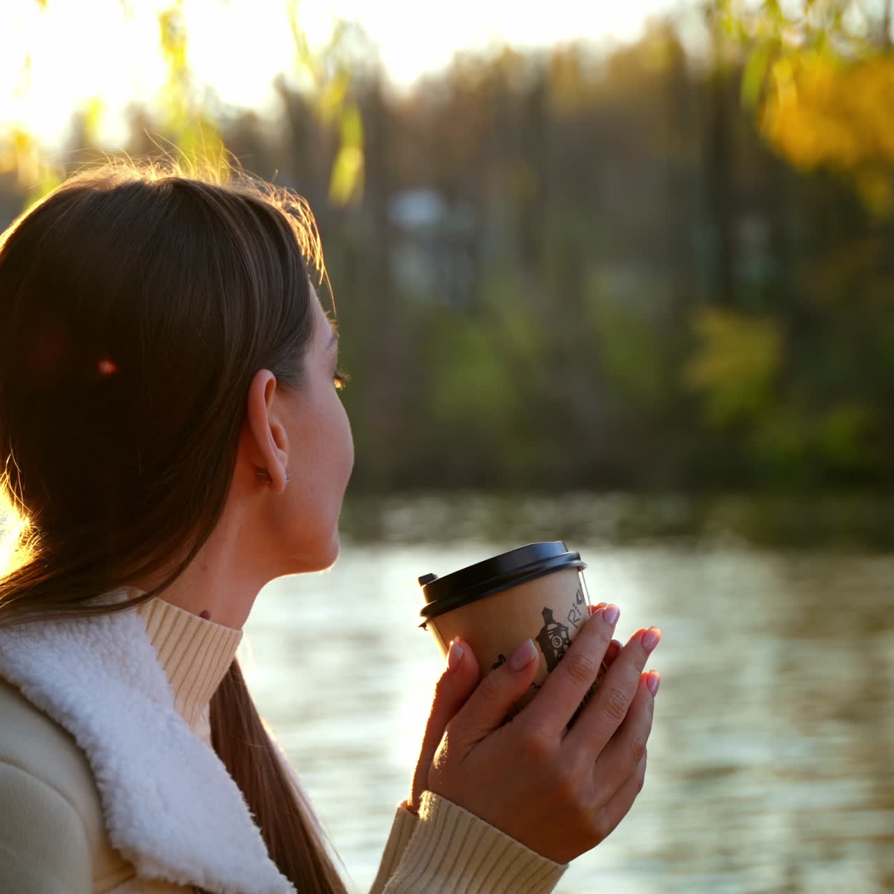 Young Caucasian lady having drink at the river bank. Woman enjoys beautiful autumn day and looking at the river. Blurred backdrop