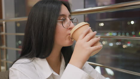 close up of young fair skinned woman wearing glasses sipping juice from paper cup while gazing thoughtfully outside through glass panel with blurred city lights