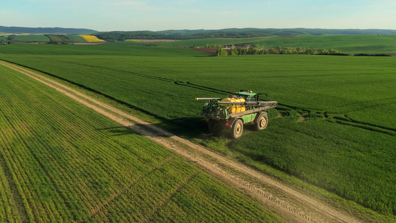 Vista aerea con drone di un trattore irroratore su una strada sterrata che irrora vasti campi verdi di grano invernale con colline ondulate sullo sfondo, Moravia, Repubblica Ceca