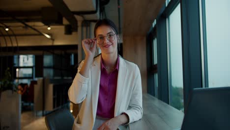 Portrait-of-a-brunette-girl-in-round-glasses,-a-white-jacket-and-a-pink-shirt-who-smiles-and-looks-at-the-camera-near-a-panoramic-window-in-a-modern-office