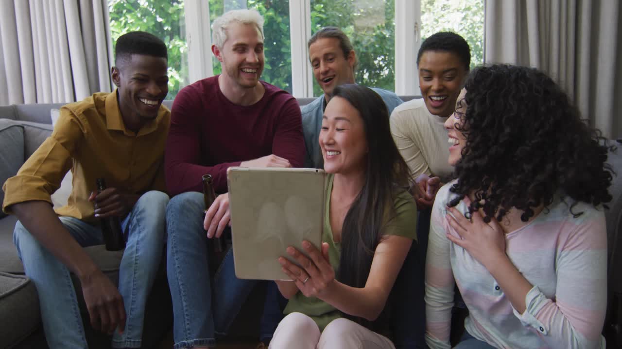 Diverse group of happy male and female friends looking at tablet and laughing in living room