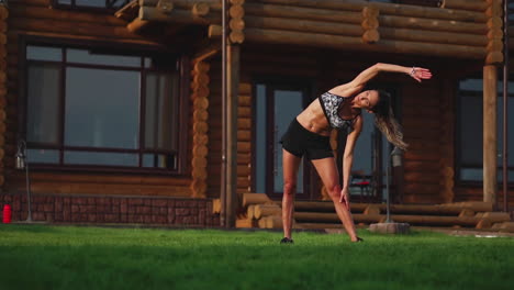 A-slim-and-beautiful-woman-in-sports-clothes-with-an-open-press-is-preparing-to-start-training-on-the-lawn-near-her-house-Performing-slopes-flexing-back-muscles