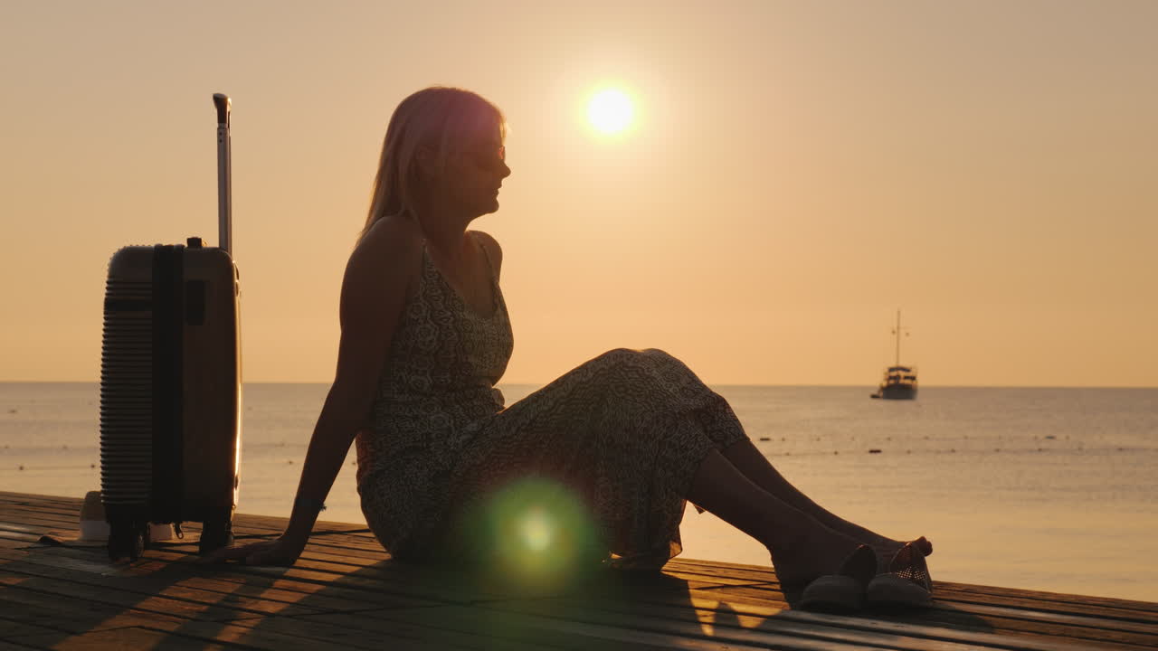 una mujer adulta se sienta en un sarafan de verano en la playa se sienta en un banco y sostiene una maleta en wh