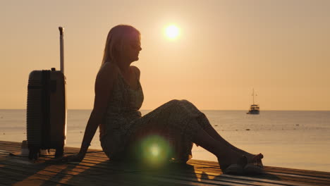 An-Adult-Woman-Sits-In-A-Summer-Sarafan-On-The-Beach-Sits-On-A-Bench-And-Holds-Onto-A-Suitcase-On-Wh