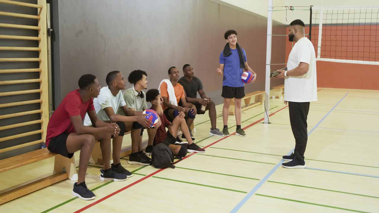 Vielfältiges Männerteam hält Volleybälle auf der Bank in der Turnhalle und reagiert auf den Trainer, der Spieler zum Training ruft.