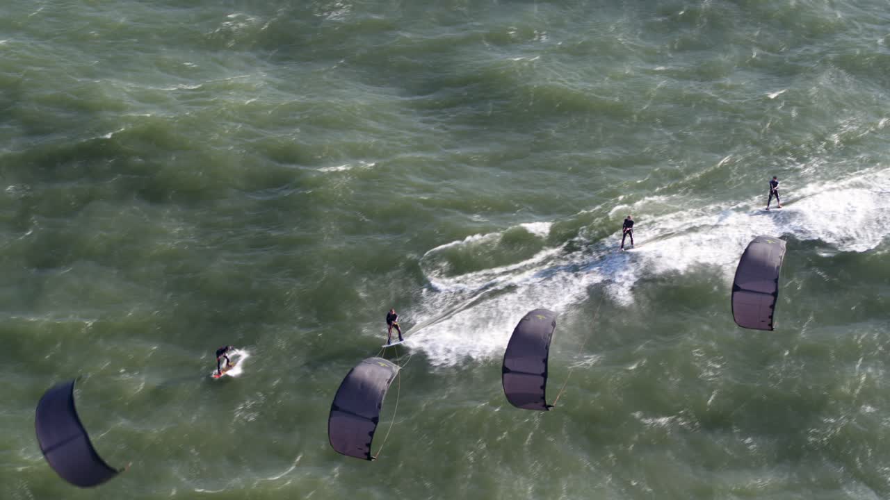Aerial wide shot of four kite surfers racing across rough ocean swells jumping and doing stunts