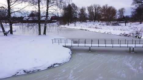 Small-city-park-with-bridge-over-frozen-lake-water-in-winter-season,-aerial-view