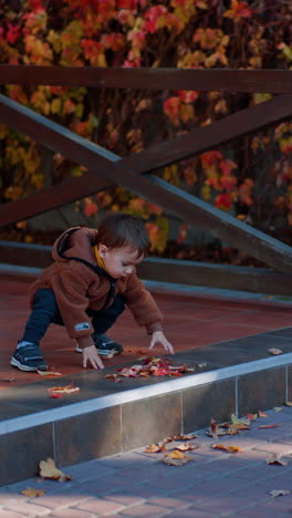 adorable caucasian toddler playing with dry autumn leaves outdoors. lovely kid throwing leaves and jumps off the porch. vertical video