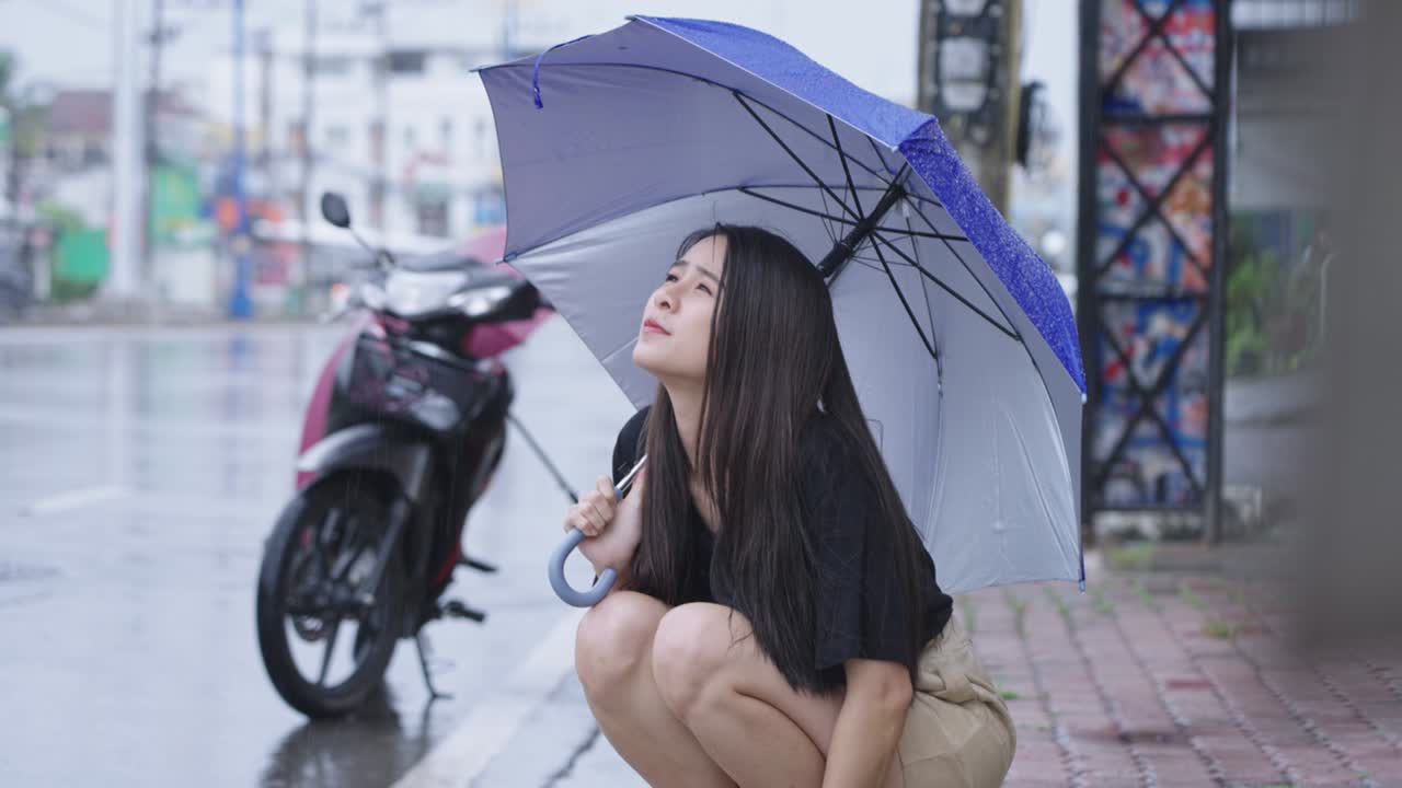 우산을 들고 있는 아름다운 아시아 소녀가 길가 에 앉아 비가 오는 날, 비가 오는 계절에 걸어가며 손을 내밀고 비방울을 만지며, 오토바이가 고장 났고, 비에 혀 실망감을 느다.