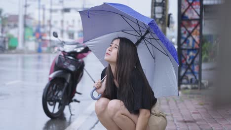 a pretty asian girl holding umbrella sit down on street side walk on raining day, rainy season reach her hand out touching rain drops, broke down motorbike, feeling disappointed stuck in the rain