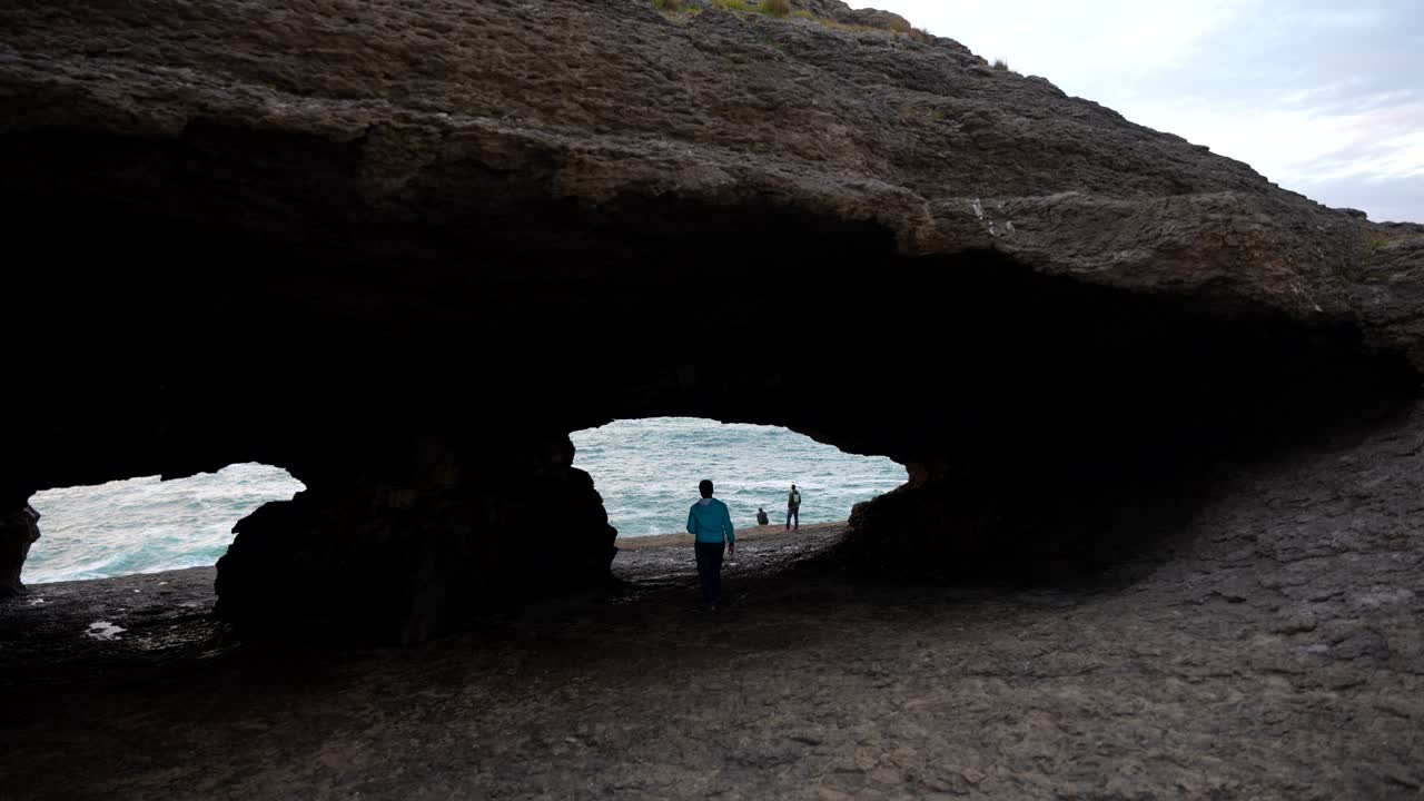 Man walking through La Ojerada Cave to join two others by the sea near a rocky cliff in Cantabria