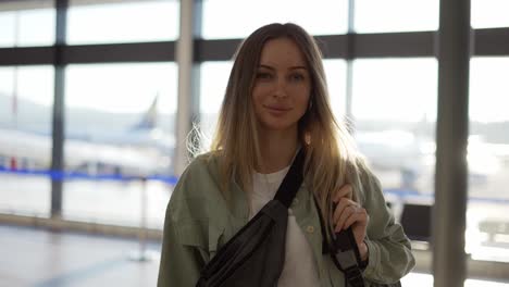 portrait of smiling young blonde girl traveller walks with backpack by airport terminal