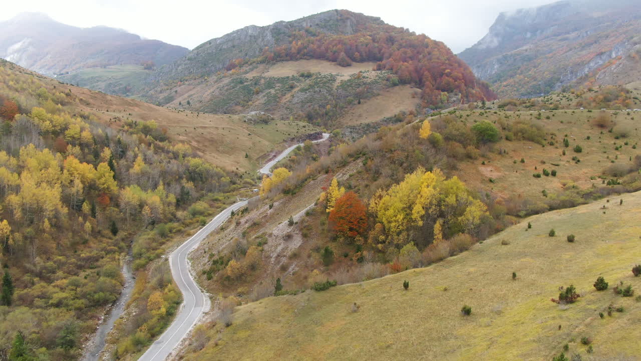 A winding road cuts through hills covered in trees Some of the trees have leaves changing color There are mountains in the distance