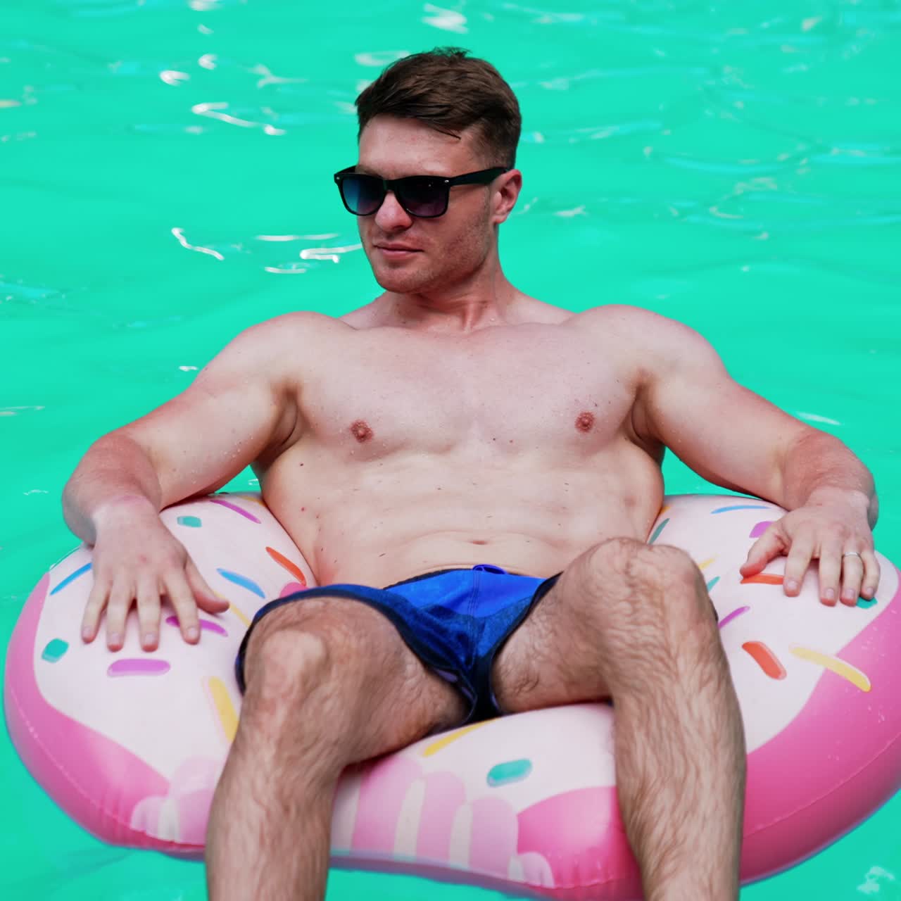 Vacation time in the city. Handsome Caucasian athletic man wearing sunglasses swims sitting in the inflatable donut