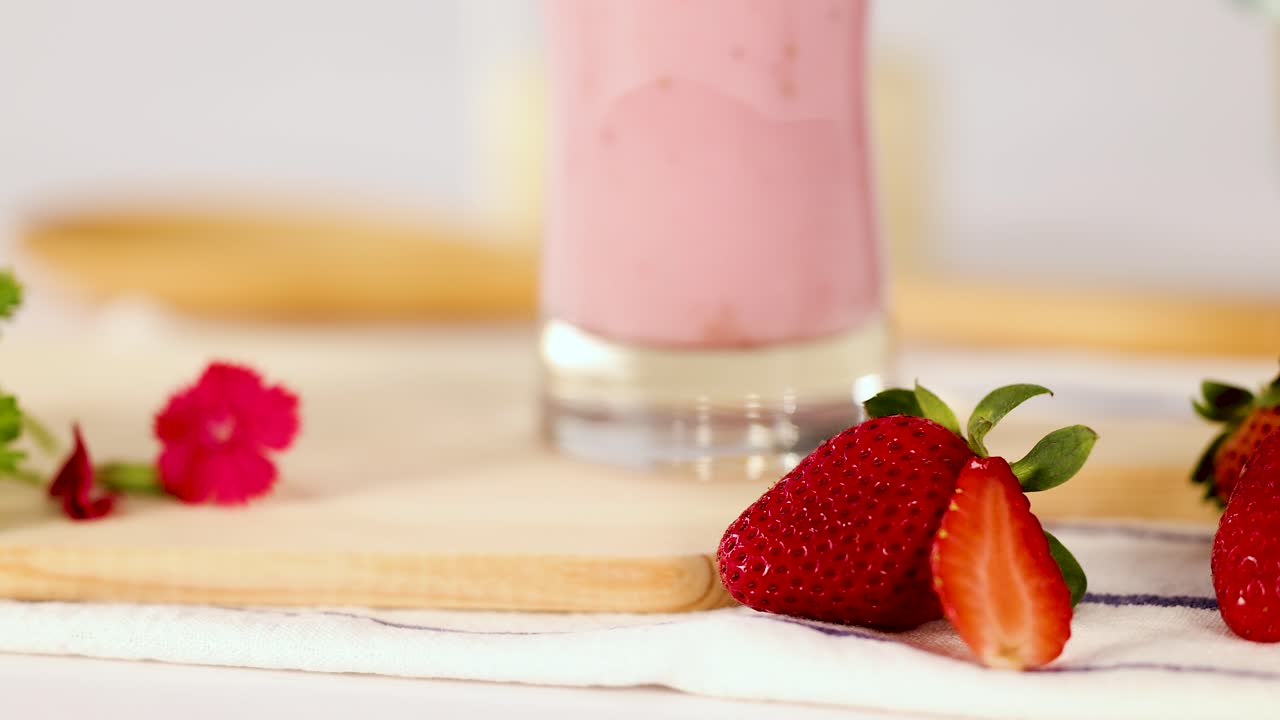 Frullato rosa alla fragola in un bicchiere con fragole, fiori e luce diurna soffusa, la telecamera si sposta lentamente lateralmente